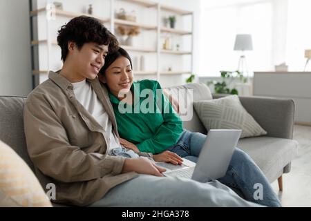 Weekend pastimes concept. Married young Asian couple using laptop at home, relaxing on couch together, copy space Stock Photo