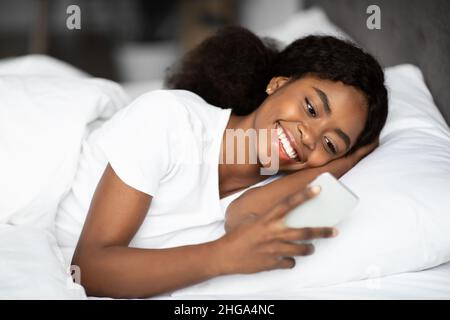 Happy young black lady laying in bed with smartphone Stock Photo