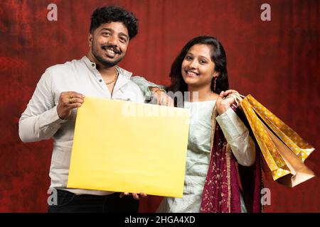 young Indian couples with empty sign board and shopping bag in hand looking at camera on studio background - promotional fetive offfers and deals Stock Photo