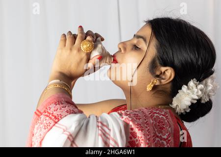 A beautiful Indian woman in red saree blowing conch shell on white background Stock Photo