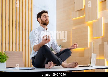 Businessman with elegant suit sitting at computer desk in the office meditating - Adult man doing yoga meditation and deep breathing at work to calm a Stock Photo