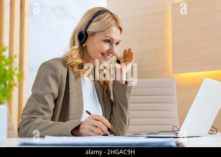 Businesswoman with elegant suit sitting at computer desk in the office and joining in a video conference - Mature adult woman working at laptop and te Stock Photo
