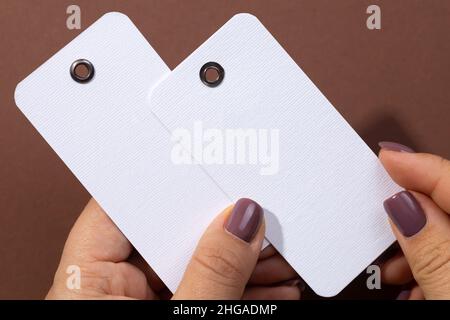 Two white blank tags of rectangular shape made of cardboard with small holes in upper part for clothes in female hands in center of brown background Stock Photo