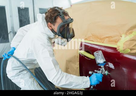 Auto painter in a protective suit and mask sprays a thinner or other cleaner on a car door using a spray gun. Preparation of vehicle body elements for Stock Photo