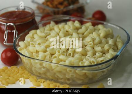 Boiled macaroni pasta inside a square glass bowl. Shot on white background with tomato red sauce in a jar and raw macaroni pastas around on the table. Stock Photo