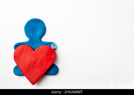 A blue plasticine man holds a red plasticine heart in his hands on a white background, flat lay, copy space. Heart medicine concept. Valentine's day c Stock Photo