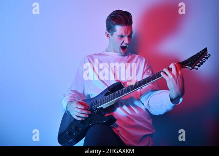 Portrait of young man artist singing, screaming, playing electric guitar Hobby, music concept. Rock Star The guy in neon plays the guitar and sings. M Stock Photo