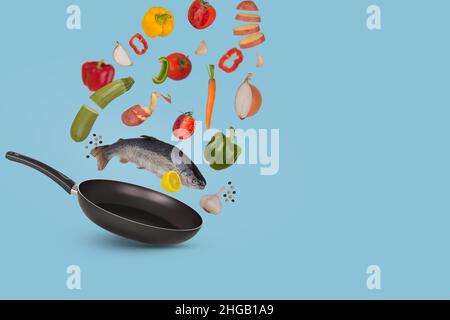 Creative food idea made of raw whole trout fish with different vegetables and lemon flying above frying pan for tasty cooking, isolated on blue backgr Stock Photo