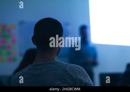 Back view of students during class during lecture. Professor next to a projected screen on the wall of a modern classroom. Stock Photo