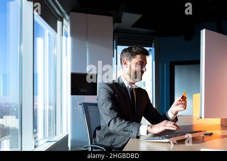 cheerful bearded businessman shopping online working in a modern office computer, man holding a bank credit card Stock Photo