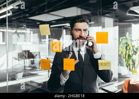 businessman in reflection at a glass task board, a man talking on the phone, hanging plans on colored paper stickers, plans for the near future Stock Photo