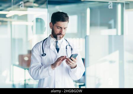 Male doctor in a white coat in a modern clinic reads information from a mobile phone corresponds with colleagues happy and smiles Stock Photo