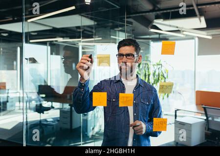 Young bearded executive manager businessman CEO writes his strategy ideas on notes on a blackboard, leading the presentation of a draft business plan Stock Photo