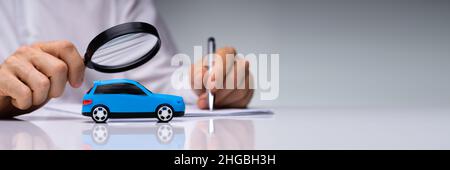Person Scrutinizing A Car Model Using Magnifying Glass On Desk Stock Photo