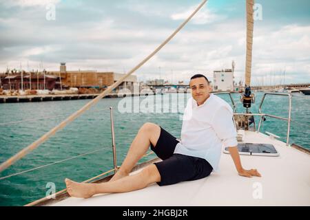 A man sits on the bow of a sailing yacht and enjoys life. Handsome guy in a white shirt, businessman, freelancer resting in the sea or ocean. Stock Photo