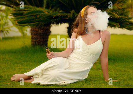 A vape woman on vacation, under a palm tree. Rest, smoking, nicotine, pleasure, danger. Vape liquid. Vaping THC e cigarette. Stock Photo