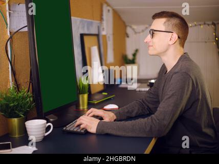 Stay at home, a man works at home. Freelance, a man studying a new program, online. Vertical computer, green background. A man with glasses sits at ho Stock Photo