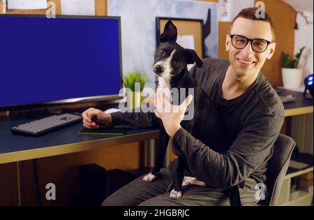 The dog sits in the man’s hands. A man works at home, a graphic designer, a creator works at home on freelance with a dog. The pet makes it difficult Stock Photo