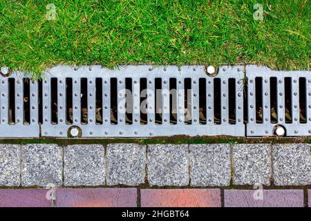 gray grating of the drainage system for drainage of rainwater in the park at the edge of the sidewalk from a stone tile with a green lawn, landscaping Stock Photo