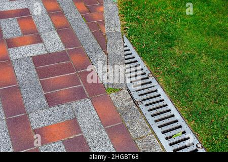 drainage grate bolted to storm drain at corner of pedestrian pavement walk path made of stone brick tiles with pattern in backyard with green grass cl Stock Photo