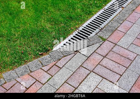 drainage grate bolted to storm drain at corner of pedestrian pavement walk way made of stone brick tiles gray and red with pattern in backyard with gr Stock Photo