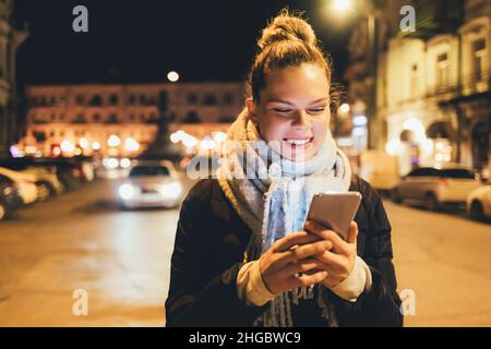 Happy young woman using her smartphone while standing on city street near road with cars on winter night Stock Photo