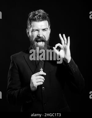 Young man wearing priest uniform standing over white background doing ok gesture with hand smiling, eye looking through fingers with happy face. Stock Photo