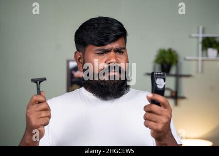 young beard man confused or thinking about what to use between razor blade and trimmer or shaver machine at home - concept of grooming, self care and Stock Photo