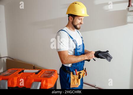 Professional electrician technician in uniform and hard hat putting on protective gloves while standing indoors Stock Photo
