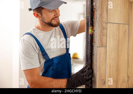 Smiling handyman, male worker in uniform using spirit level while checking measurements of the wall indoors Stock Photo