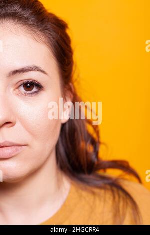 Close up of young woman standing in photography studio, with half of face looking at camera to pose for pictures. Caucasian happy person standing over isolated orange background. Stock Photo
