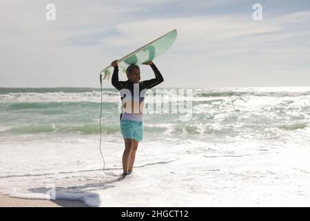 Biracial senior man carrying surfboard over head while standing on shore at sunny beach against sky Stock Photo