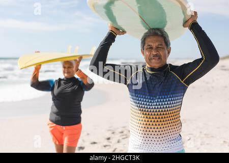 Multiracial senior couple enjoying retirement together while carrying surfboards over heads at beach Stock Photo