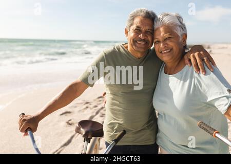 Portrait of smiling senior multiracial couple with arm around bicycles at beach on sunny day Stock Photo