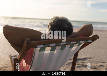 Biracial senior man relaxing with hands behind head sitting on folding chair at beach during sunset Stock Photo