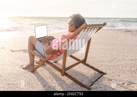 Biracial senior man sitting on chair using blank screen laptop with copy space at beach Stock Photo