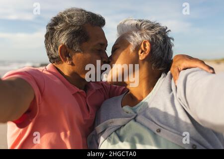 Romantic senior multiracial couple kissing each other while taking selfie at beach Stock Photo