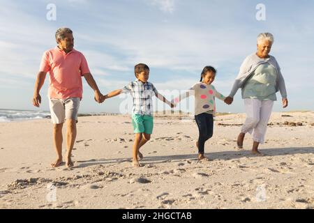 Full length of senior biracial man and woman holding hands with grandchildren while walking at beach Stock Photo