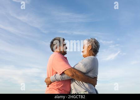 Low angle side view of smiling biracial senior couple looking while embracing each other against sky Stock Photo