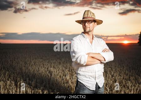 Farmer in a straw hat and glasses standing in a ripe wheat field before sunset. Agriculture production concept Stock Photo