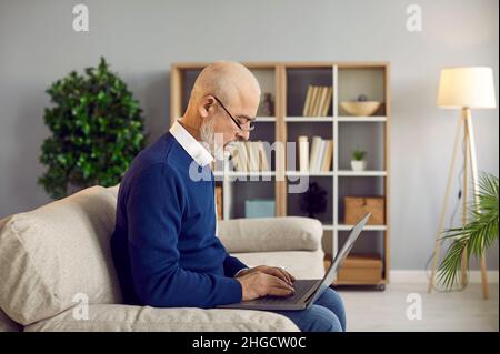 Serious senior man in glasses sitting on sofa at home and working on his laptop computer Stock Photo