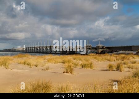 View across the sea to the Eastern Scheldt Storm Surge Barrier in Zeeland with a stormy sky, dramatic sunlight with sand dunes, grass and beach in for Stock Photo