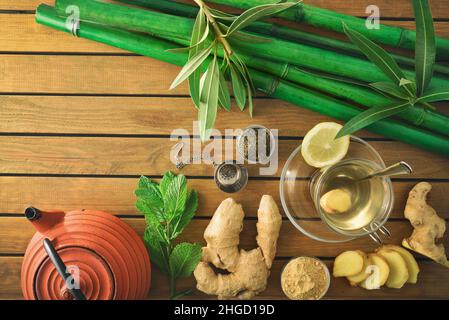 Cup of green tea with ginger mint and lemon on wooden table with leaves, metal teapot and bamboo canes on wooden table. Top view. Stock Photo