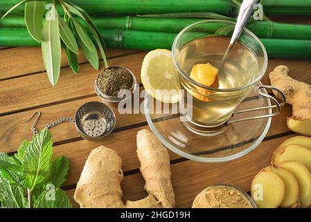 Cup of green tea with ginger mint and lemon on wooden table with leaves, metal teapot and bamboo canes on wooden table. Elevated view. Stock Photo