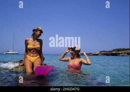 2 young women sit on rock in front seaside talking into seawater Stock Photo