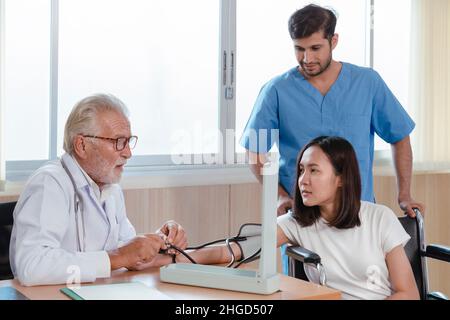caucasian senior doctor using sphygmomanometer checking blood pressure and consults to young asian patient woman in the hospital. healthcare and medic Stock Photo