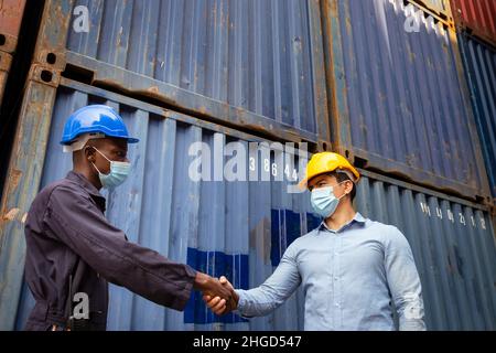 african worker and caucasian businessman wearing face mask to protect from covid or coronavirus are shaking hand in cargo warehouse shipping after suc Stock Photo