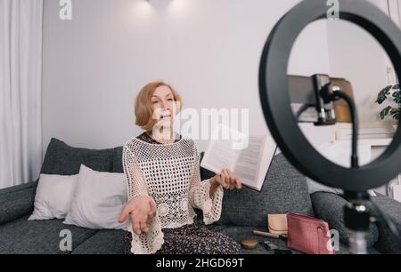 A photo of a beautiful elderly blogger woman sitting on the couch and talking about a new book in front of her subscribers, looking at a smartphone Stock Photo