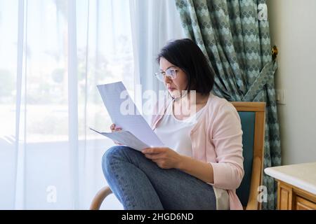 Serious mature woman reading papers documents sitting in armchair at home. Stock Photo