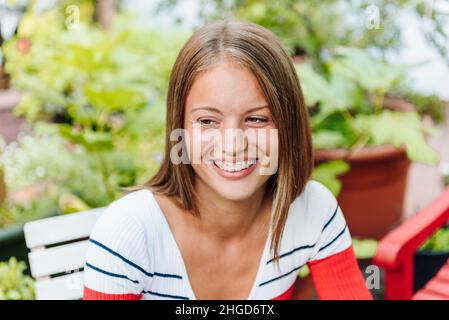 Happy young female with long hair sitting on chair near green plants on terrace of outdoor garden on summer day Stock Photo